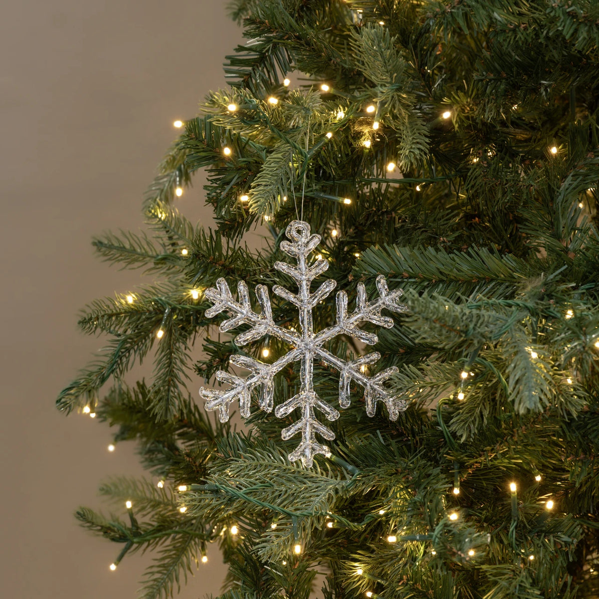 A silver glitter snowflake Christmas tree ornament hanging on a green artificial Christmas tree with warm white string lights.