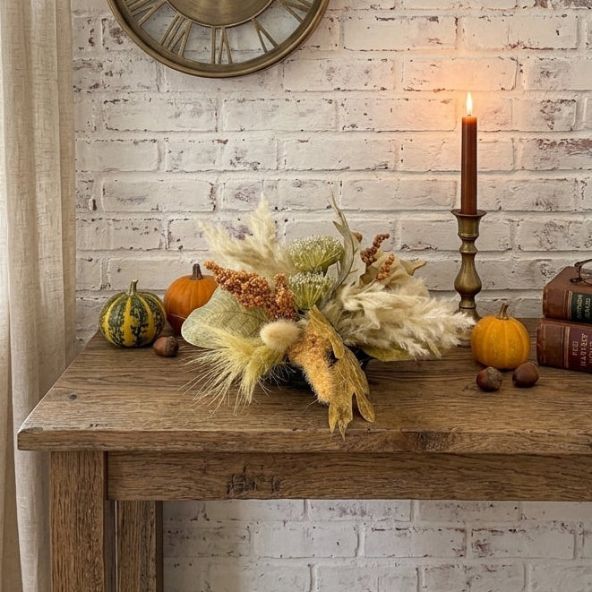An autumn-themed floral centerpiece with dried grasses, acorns, and small pumpkins displayed on a rustic wooden table against a white brick wall, with a lit candle in a brass holder.