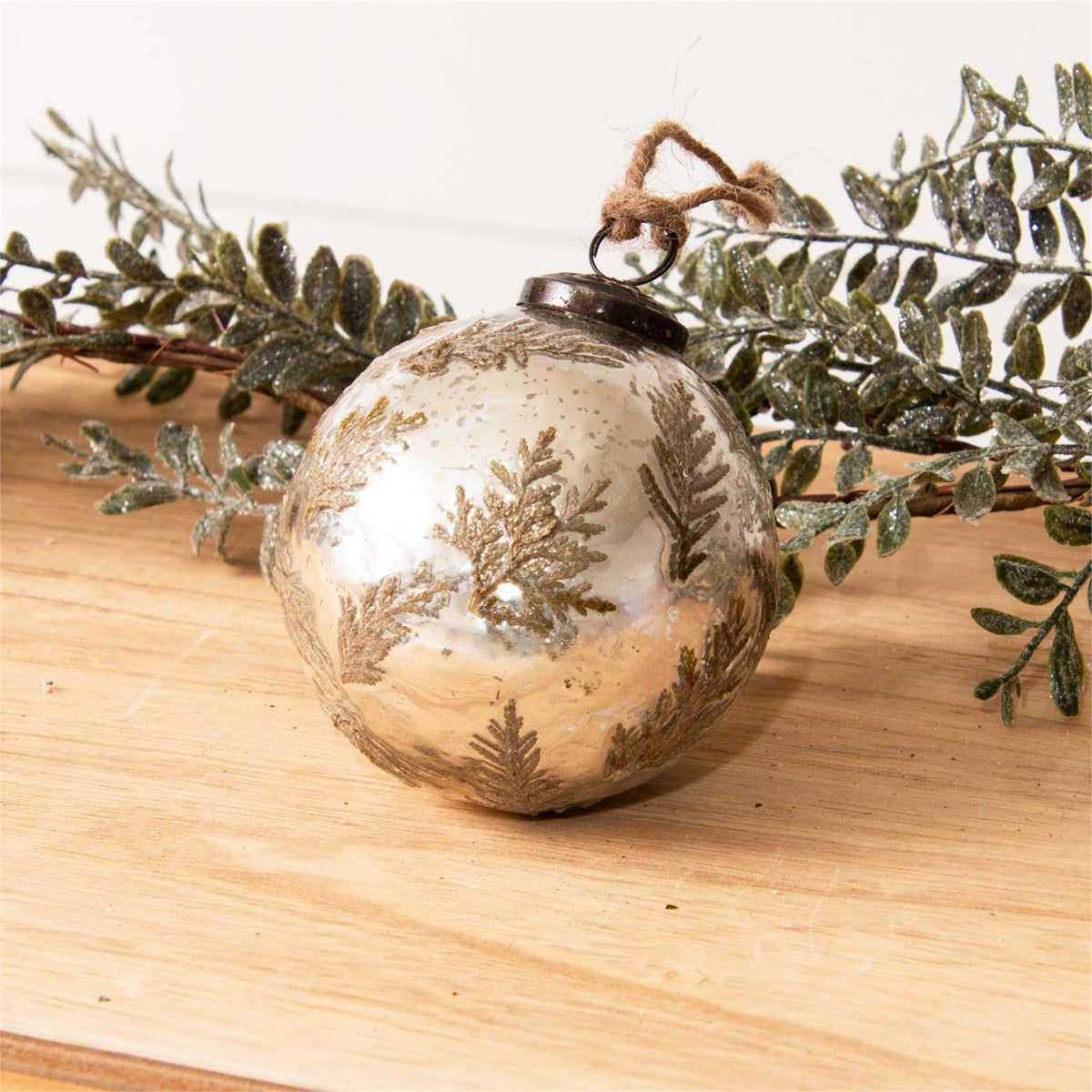 A round, metallic champagne-gold Christmas ornament decorated with gold-toned snowflakes, resting on a light wooden surface with green frosted pine branches behind it.