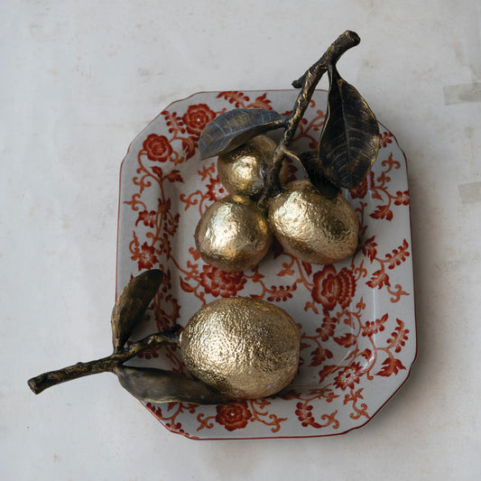 Three metallic gold decorative fruit sculptures with dark brown branches and leaves, arranged on a white ceramic tray with a red floral pattern, set against a light marble background.