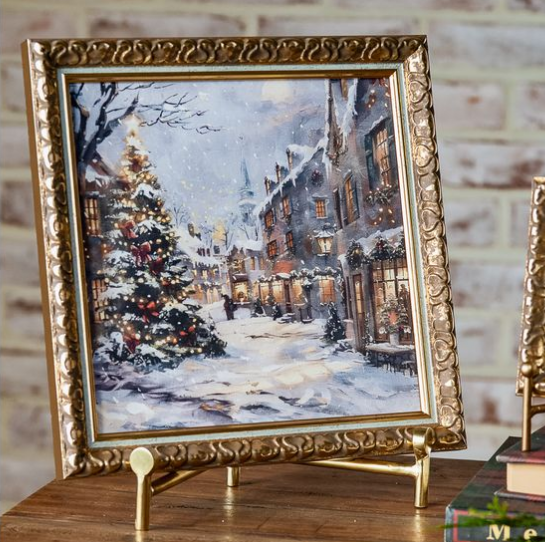 A square framed winter village painting displayed on a gold easel stand. The artwork depicts a snowy street with decorated Christmas trees, warm glowing windows, and snow-covered buildings.