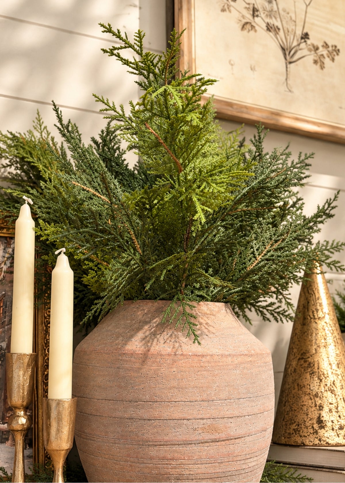 A large terracotta-colored ceramic planter filled with green evergreen branches, styled with two cream pillar candles in gold holders and a gold cone ornament against a white shiplap wall.