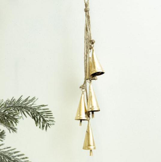 A hanging set of five small metallic gold bells suspended from a natural jute rope, shown against a plain off-white background with a pine branch entering from the left edge.