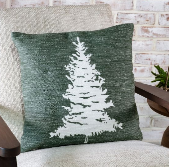 A square throw pillow with a textured dark green background and a white knit pine tree design, resting on a beige upholstered armchair in front of a white brick wall.