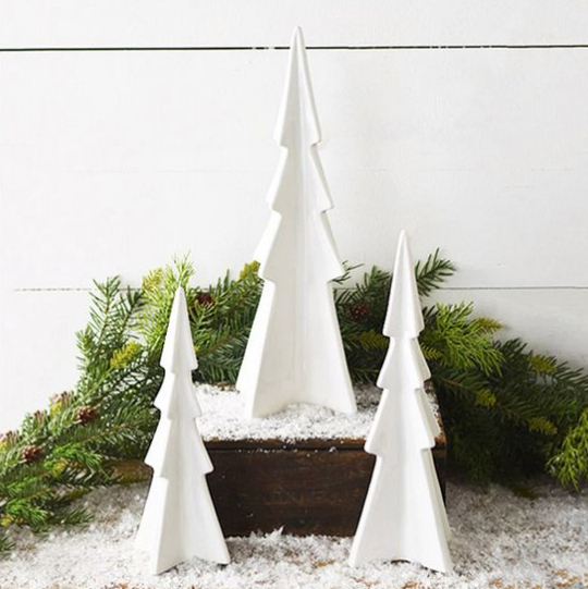 Three white geometric Christmas tree ornaments in different sizes displayed on a dark wooden box with faux snow and green pine branches against a white paneled wall.