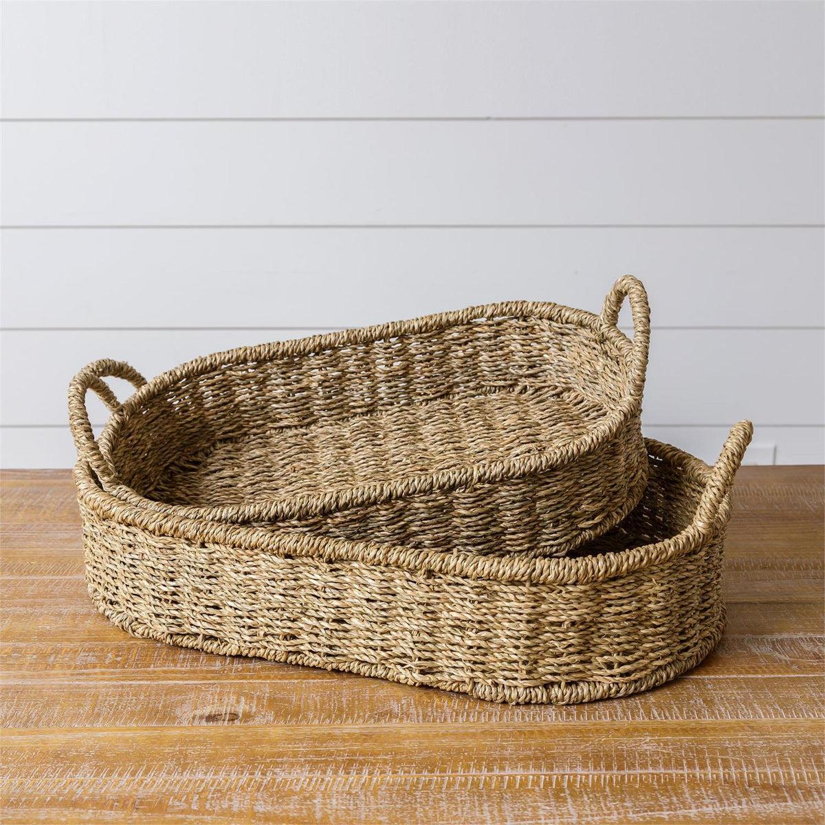 Two woven baskets with handles on a wooden surface with a white wooden panel background