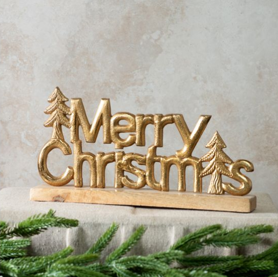 A gold-painted metal 'Merry Christmas' sign with tree cutouts, mounted on a light wood base, displayed on a stone surface with green pine branches in the foreground.