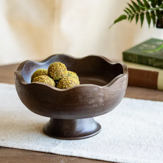 A dark brown wooden pedestal bowl with a scalloped rim, shown on a white textured runner with decorative confections inside. The background features a soft-focus warm setting with a book and a green leafy plant.