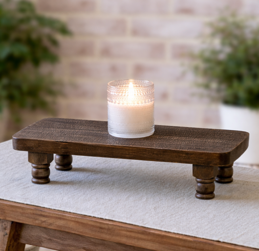 A lit white candle in a textured clear glass jar sits on a dark brown wooden elevated tray with turned legs, placed on a light fabric-covered table with a blurred brick wall and greenery in the background.