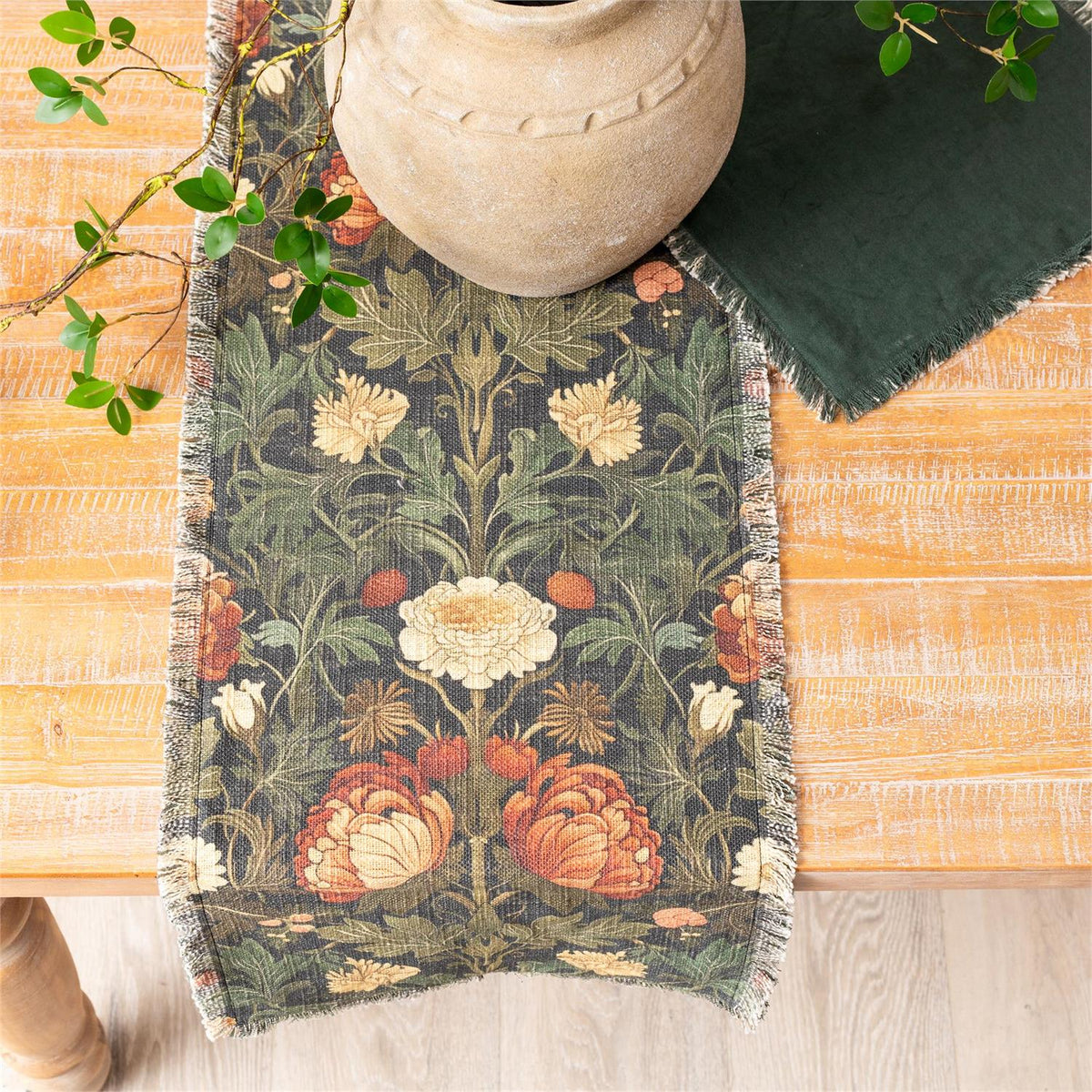 Floral patterned table runner on a wooden table with a vase and green fabric.