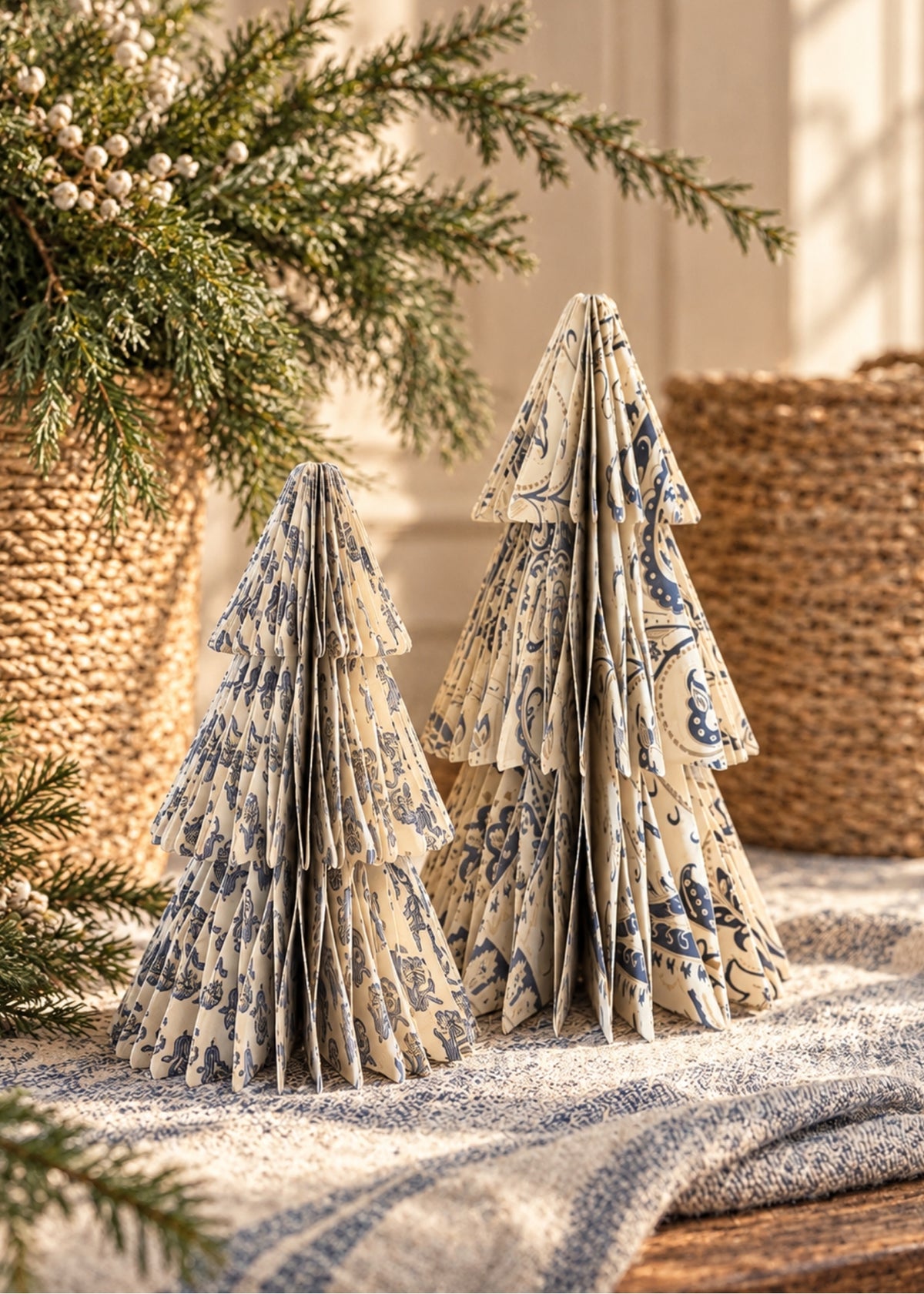 Two decorative paper Christmas trees with a blue toile pattern on a cream background, styled on a textured blanket with pine branches and woven baskets.