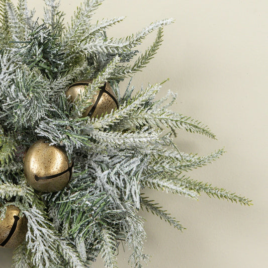 Frosted green pine branches with white snow-like dusting and several metallic gold jingle bells, photographed against a plain warm beige background.