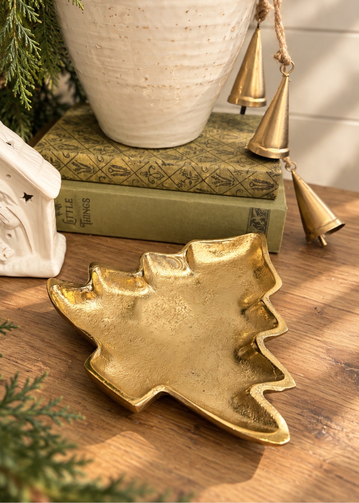 A gold-toned metal Christmas tree-shaped tray styled on a wooden tabletop with books, a ceramic pot, hanging brass bells, and evergreen branches.