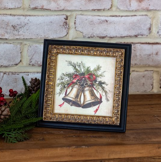 A gold and black framed Christmas art print featuring two gold bells with green pine branches and a red bow, displayed on a wooden surface against a white brick wall with holiday greenery and pinecones.