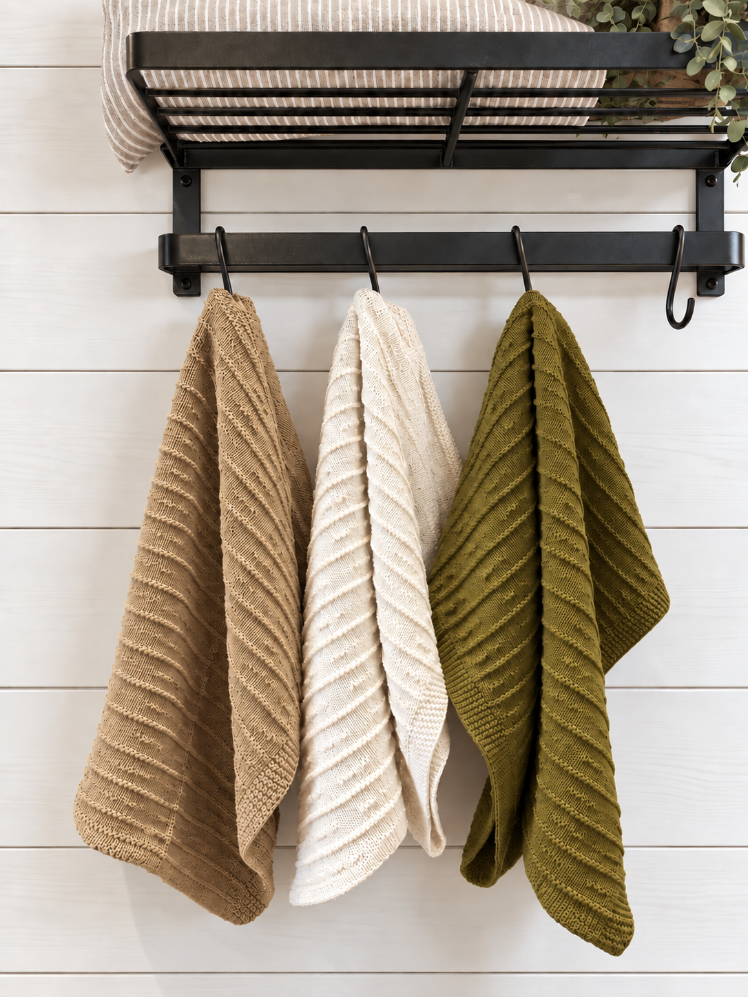 Three textured knit kitchen towels in tan, cream, and olive green hanging from a black metal rack against a white wood plank wall.