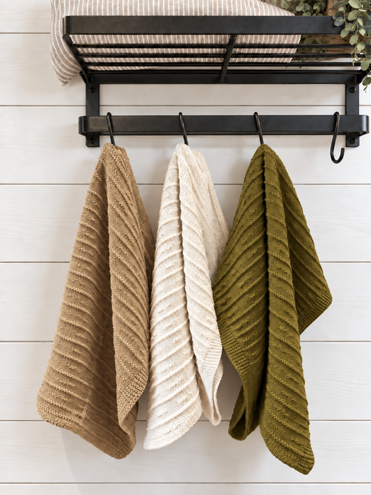 Three textured knit kitchen towels in tan, cream, and olive green hanging from a black metal rack against a white wood plank wall.
