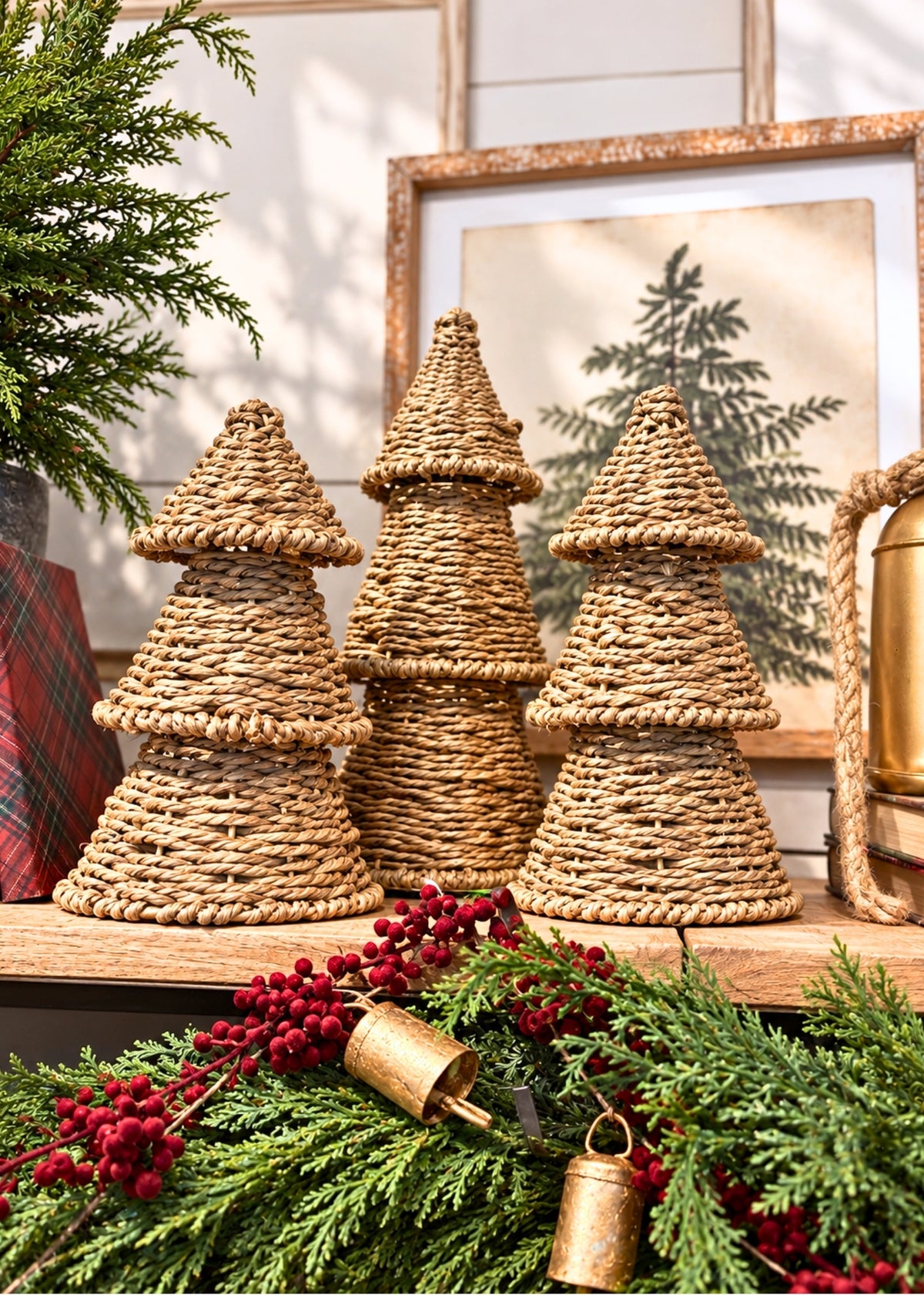 Three woven natural-fiber Christmas tree ornaments displayed on a wooden shelf, surrounded by holiday greenery, red berries, and brass bells, with a framed print in the background.