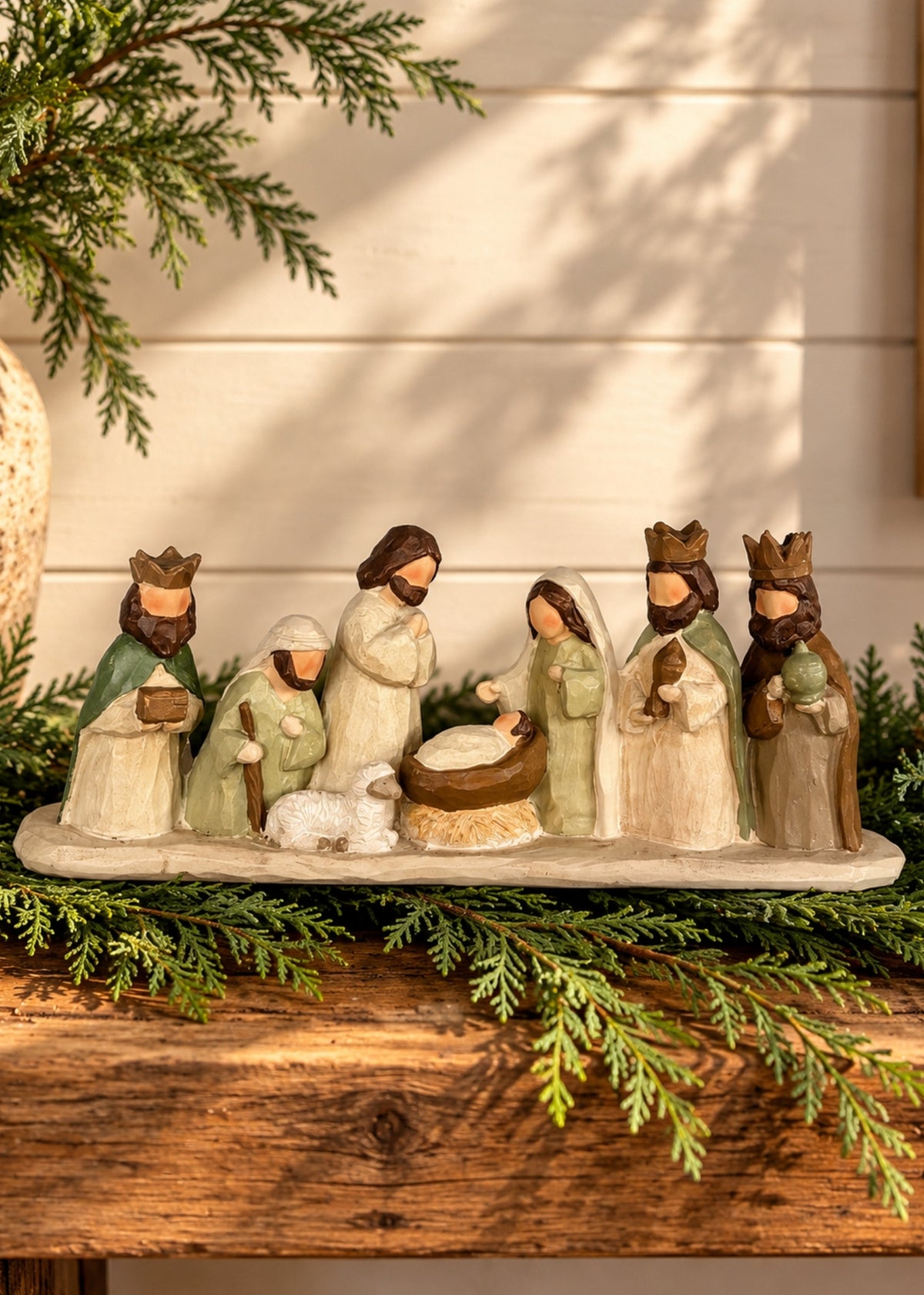 A rustic nativity scene figurine set displayed on a wooden surface with green pine branches. The set includes Mary, Joseph, baby Jesus in a manger, a shepherd, an angel, and three wise men, all crafted in a warm, earthy style.