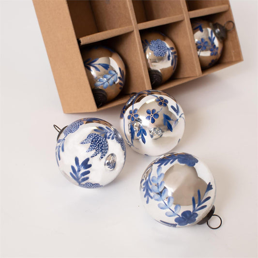 Three round glass Christmas ornaments with a white base and blue floral pattern are shown in front of a brown cardboard gift box containing more matching ornaments.