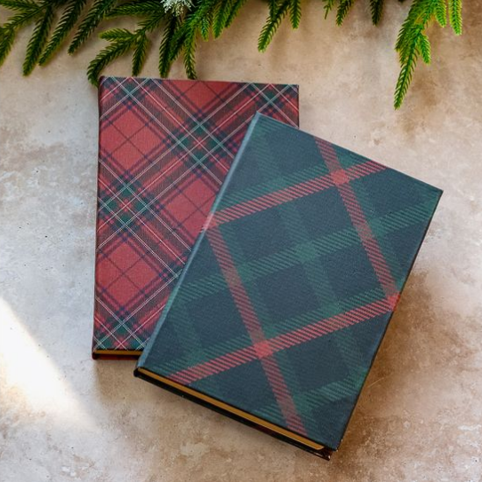 Two hardcover books with tartan plaid covers, one in red and green plaid and the other in dark green and red plaid, arranged on a light stone surface with pine branches at the top.