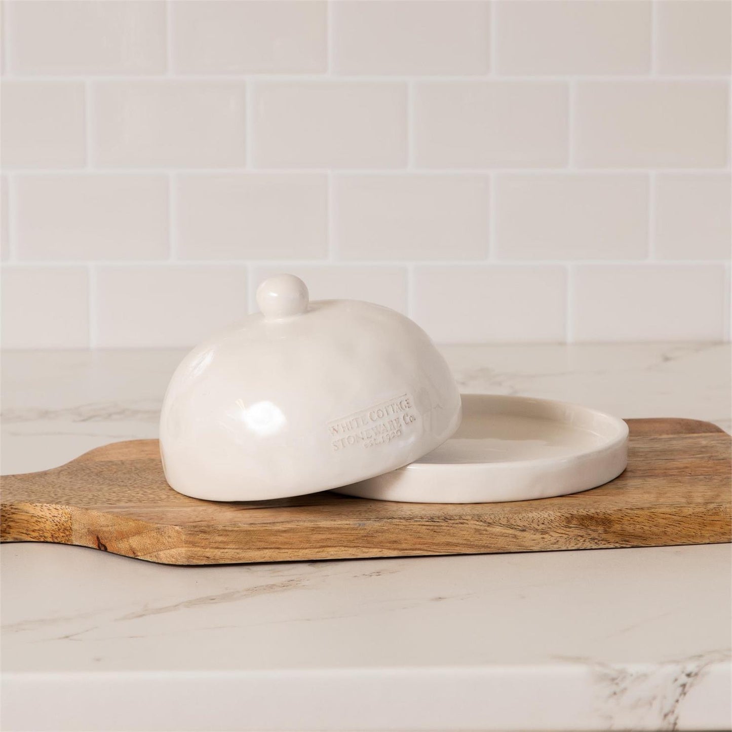 White ceramic butter dish with lid on a wooden cutting board against a tiled wall.