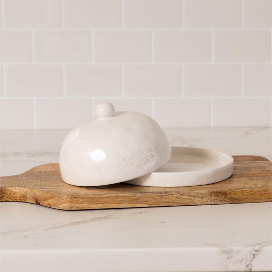 White ceramic butter dish with lid on a wooden cutting board against a tiled wall.