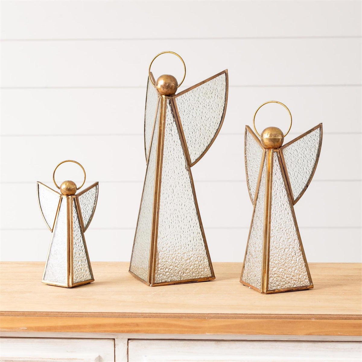 Three angel-shaped decorative lanterns with textured clear glass and gold-toned metal frames, displayed on a light wood surface against a white paneled wall.