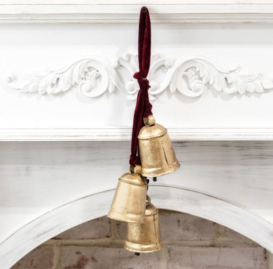 Three antique-style gold bells hanging from a dark red velvet ribbon, displayed against a white decorative fireplace mantel.