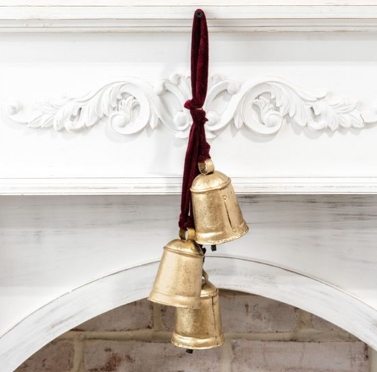 Three antique-style gold bells hanging from a dark red velvet ribbon, displayed against a white decorative fireplace mantel.