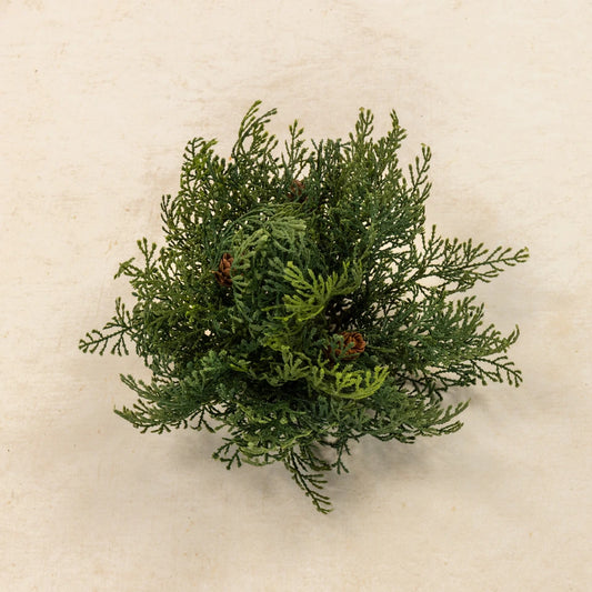 A small artificial green evergreen bush with pinecones, photographed from above on a light beige textured background.