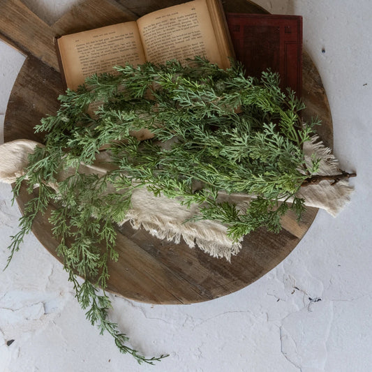 A styled flat-lay featuring a bundle of fresh green juniper branches draped over a rustic wooden board, resting on a beige linen cloth with an open vintage book and a small red book in the background.