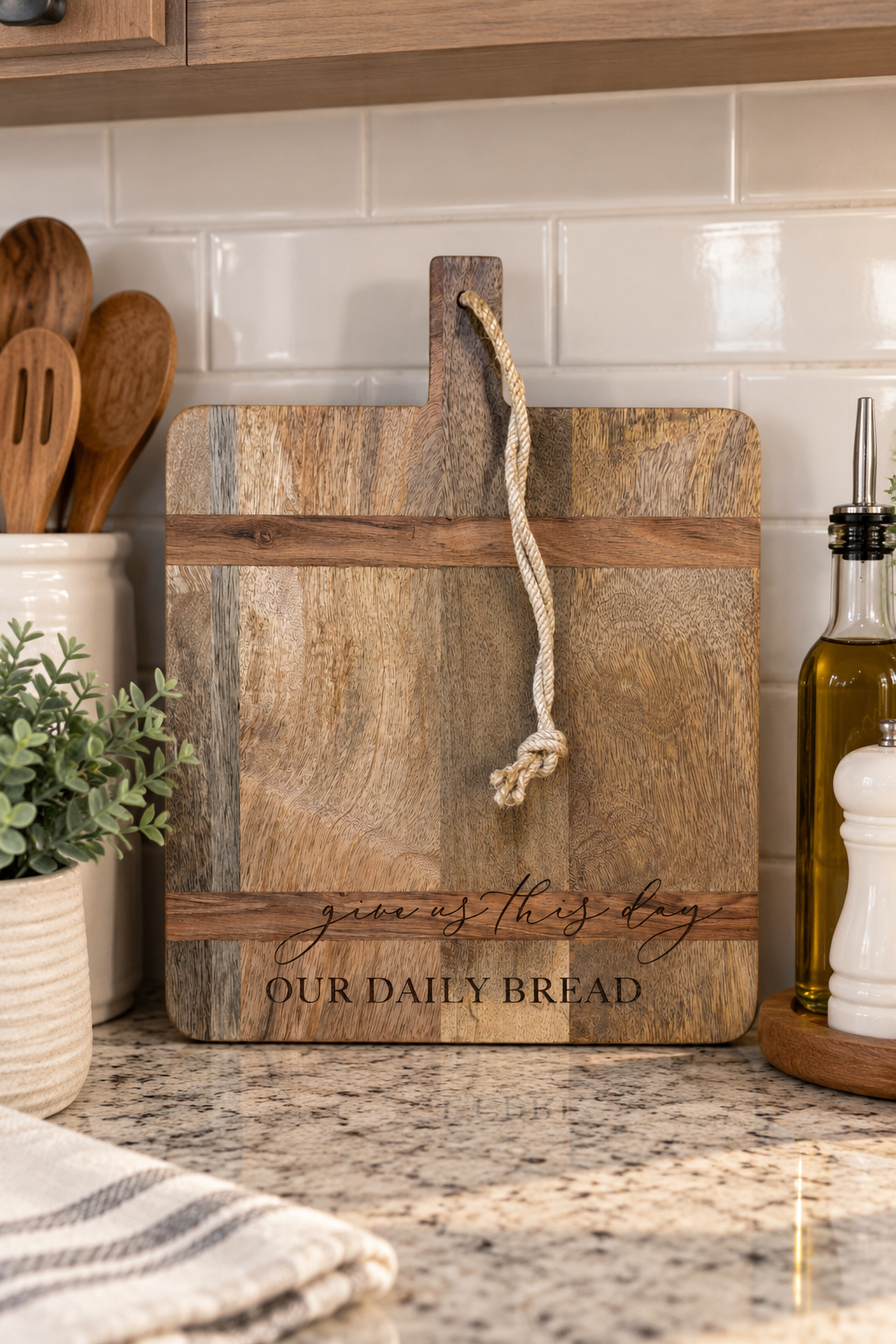 A rustic rectangular wooden cutting board with a rope handle, engraved with the words 'generous this bag OUR DAILY BREAD', displayed on a kitchen counter.