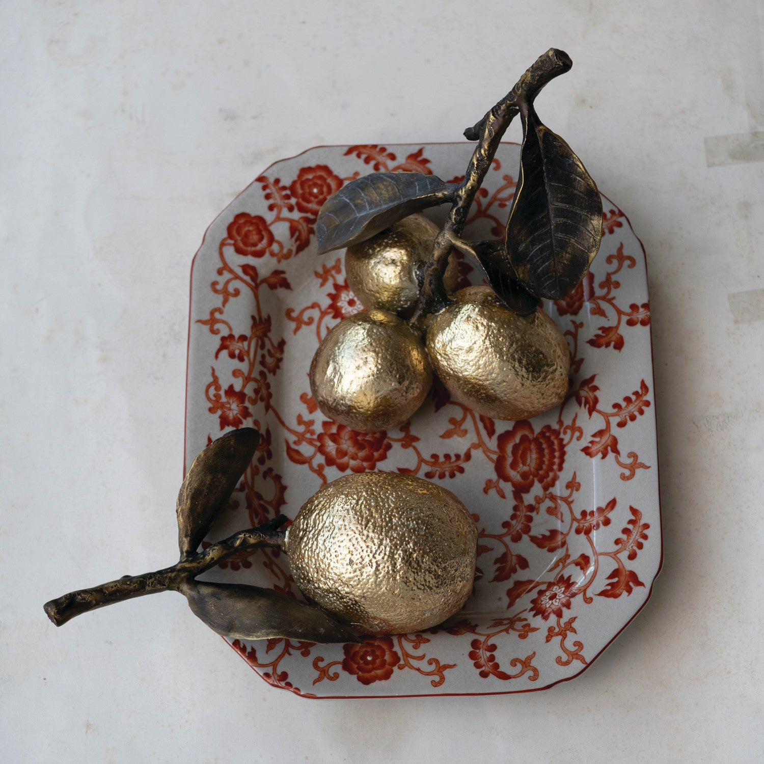 Three metallic gold decorative fruit sculptures with dark brown branches and leaves, arranged on a white ceramic tray with a red floral pattern, set against a light marble background.