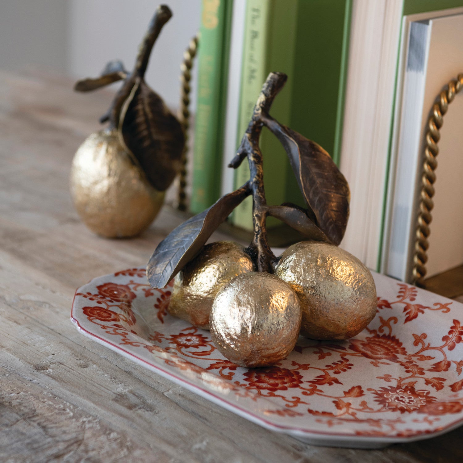 Three decorative metallic gold lemons on stem with dark brown leaves arranged on a white ceramic plate with a red floral pattern, set on a wooden surface with books in the background.