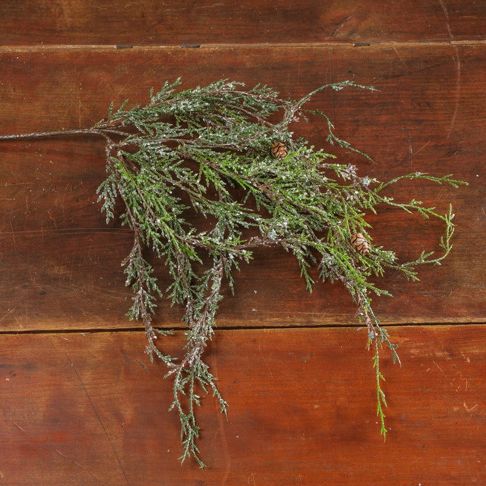 A natural-looking evergreen branch with pinecones, covered in a light dusting of white snow, laid flat on a rustic dark brown wooden surface.