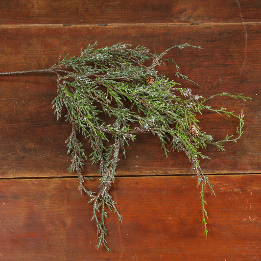 A natural-looking evergreen branch with pinecones, covered in a light dusting of white snow, laid flat on a rustic dark brown wooden surface.