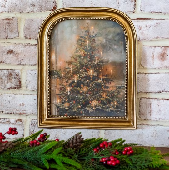 A framed vintage-style Christmas painting featuring a glowing decorated tree, hung on a white brick wall above a holiday garland with red berries and pinecones.