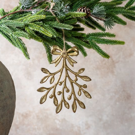 A gold metal mistletoe ornament hanging from a green pine branch against a soft beige background.