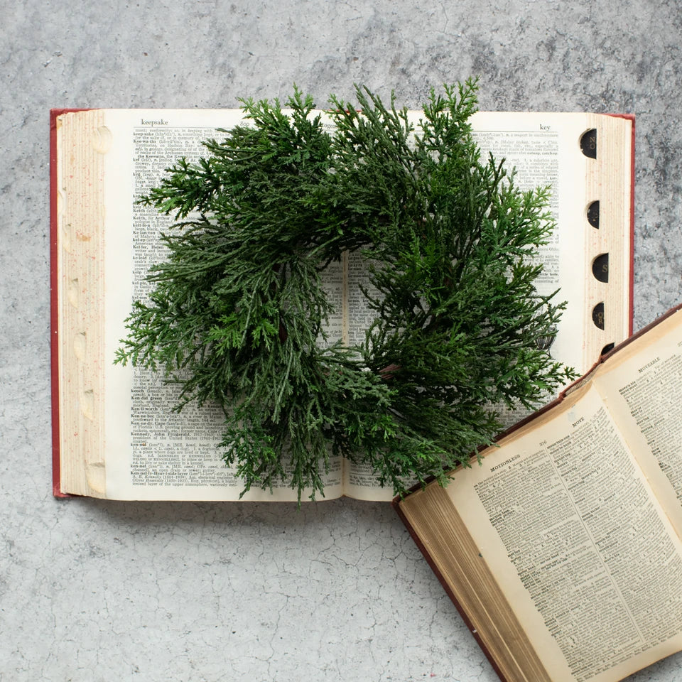 A green artificial-looking evergreen wreath placed on top of an open vintage book, with another open book at the lower right, on a light gray textured surface.