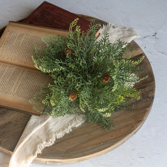A green artificial evergreen garland with small pinecones, styled on top of an open vintage book and draped over a round wooden board against a white textured background.