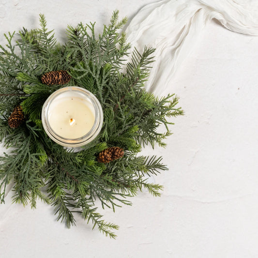 A lit glass jar candle with creamy white wax sits in the center of a green pine garland with two small pinecones, arranged on a white textured surface with a white fabric napkin in the corner.