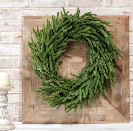 A green pine wreath with a rustic wood frame, displayed against a white brick wall background.