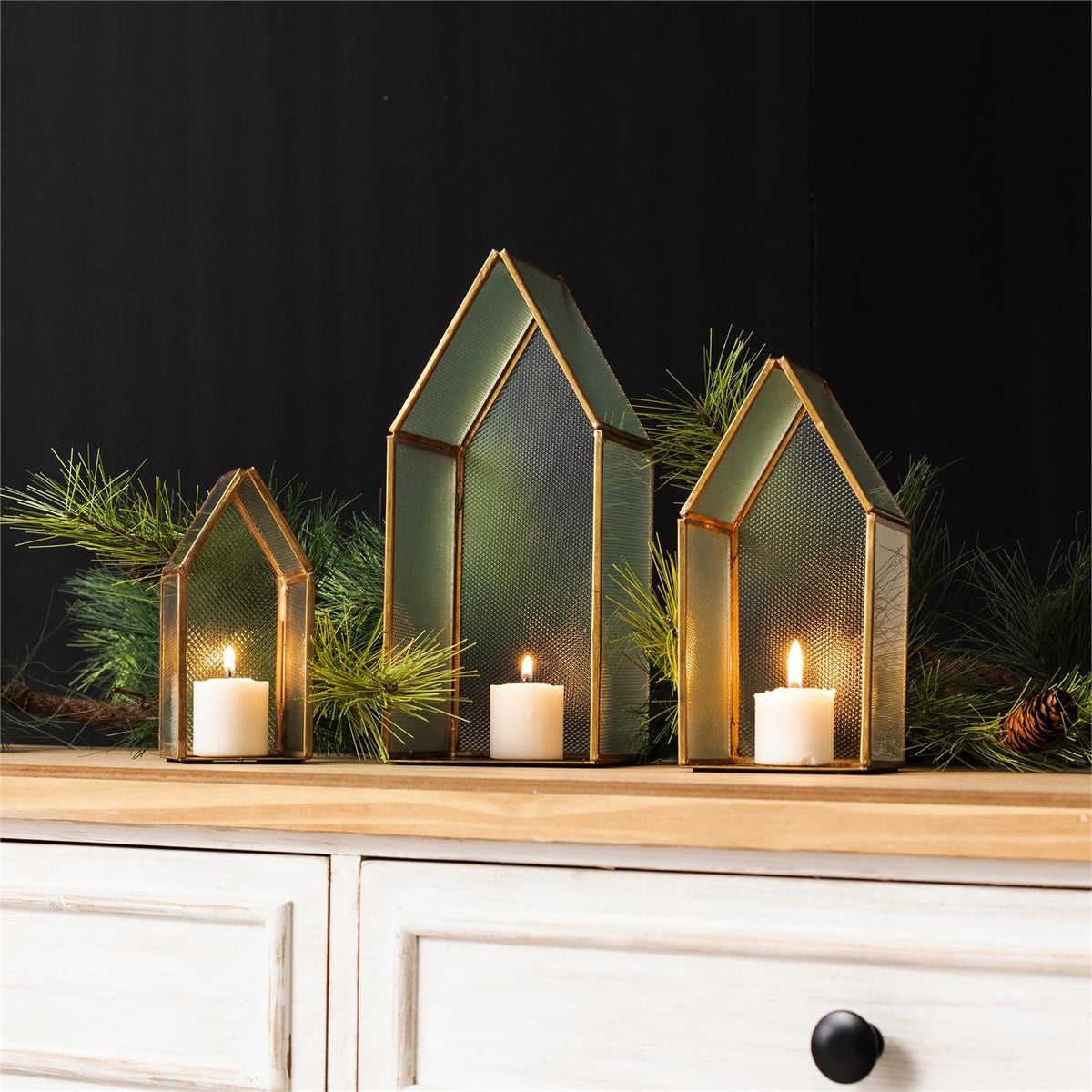 Three house-shaped glass candle holders with gold frames, each containing a white pillar candle, arranged on a wooden surface against a dark background with pine branches.