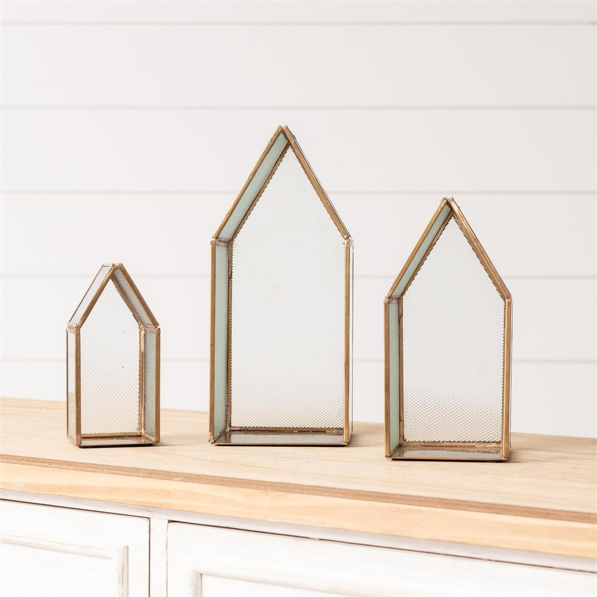 Three house-shaped glass terrariums with gold-toned metal frames, displayed in ascending sizes on a light wood surface against a white shiplap wall.