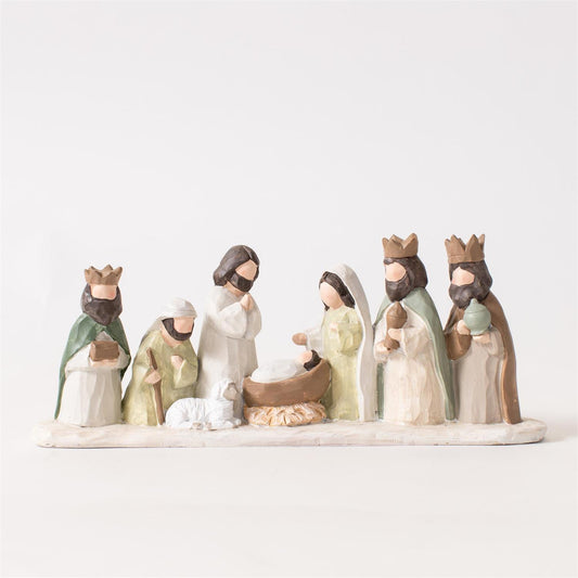A small nativity scene figurine set on a white base, showing several stylized human figures in robes around a baby Jesus in a manger, photographed against a plain light background.