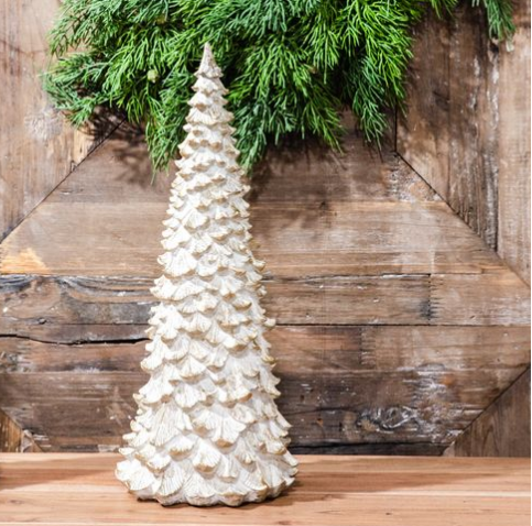 A textured, off-white resin Christmas tree centerpiece standing on a wooden surface against a rustic wood plank wall with green pine branches at the top.