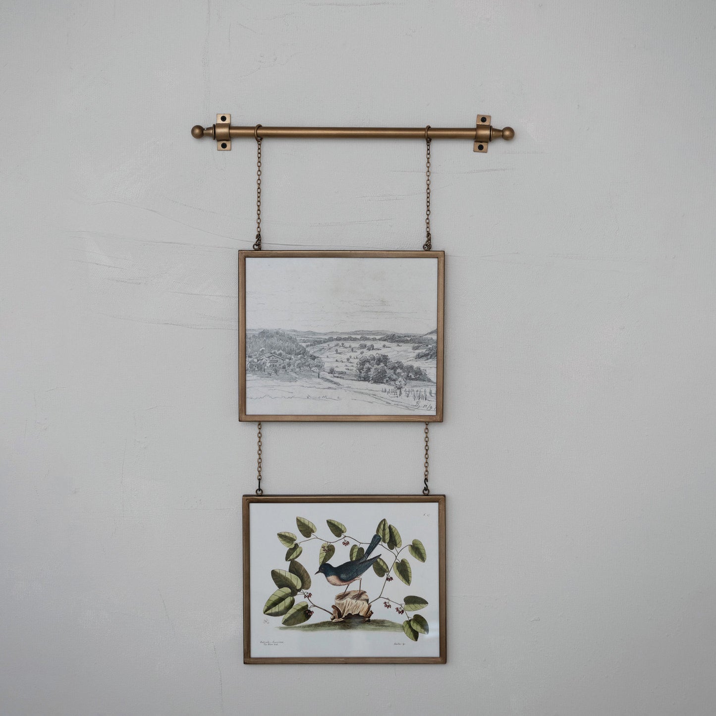 Two framed prints hanging on a wall with a decorative metal rod and chains.