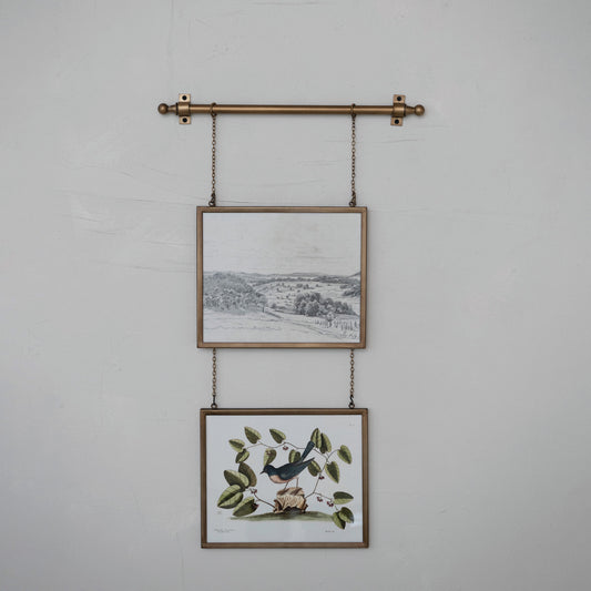 Two framed prints hanging on a wall with a decorative metal rod and chains.