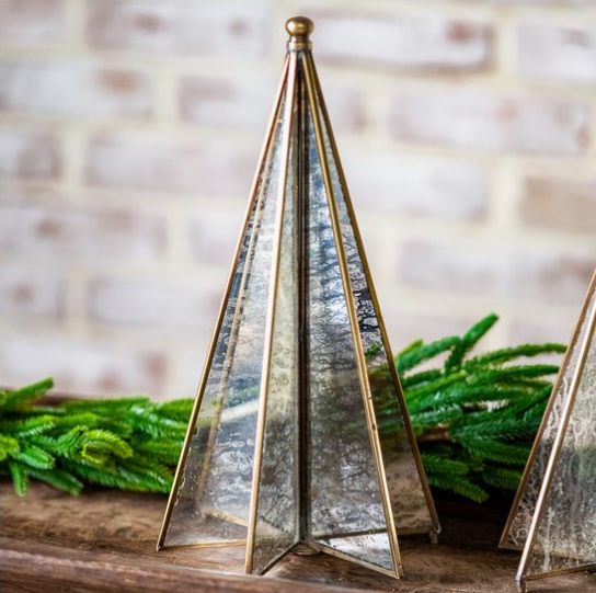 A decorative pyramid-shaped glass vase with a hammered silver finish and brass-gold metal trim, styled on a wooden surface with green pine branches in front of a soft-focus brick wall.