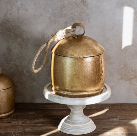 A rustic brass bell-shaped container with a rope loop handle, displayed on a white pedestal stand against a textured gray wall.